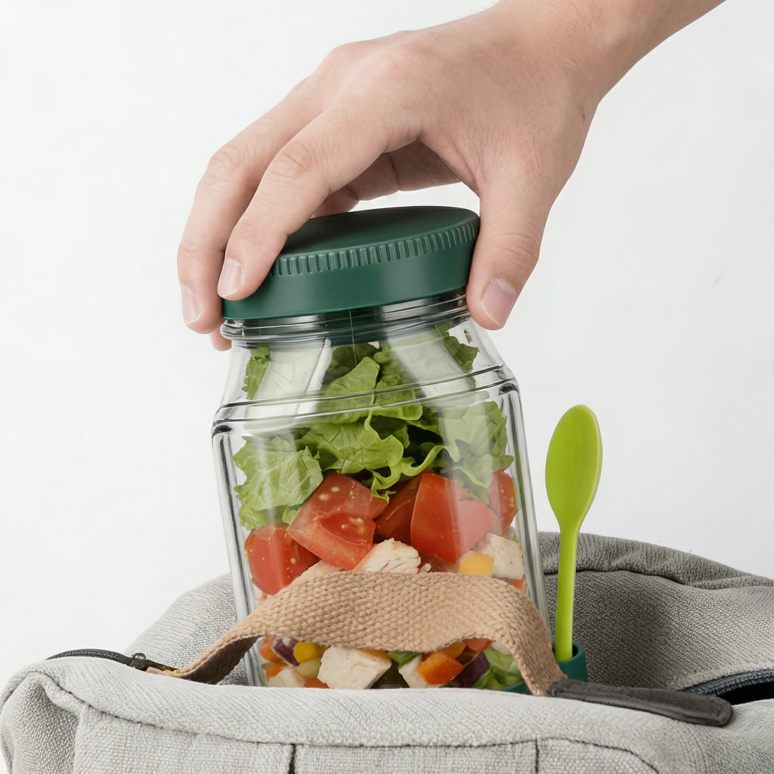 Borcan cu salată pus într-o geantă de prânz, cu capacul verde ținut în mână